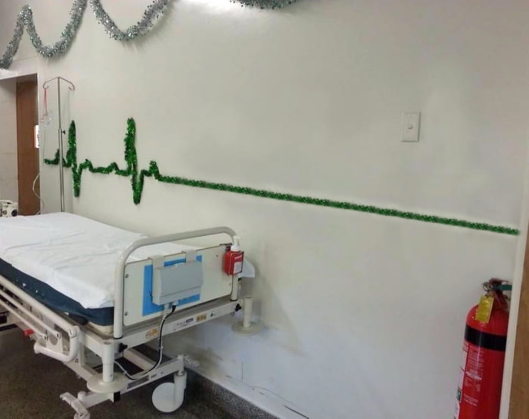 A hospital bed is next to a wall decorated with green tinsel shaped like an electrocardiogram (EKG) line, ending in a straight line. There is a fire extinguisher on the floor by the wall.