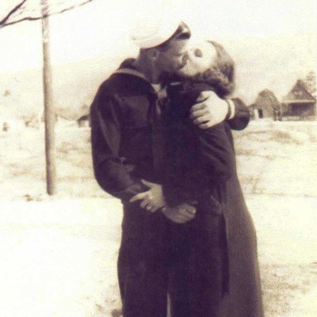 A vintage photo of a sailor in uniform kissing and embracing a woman outdoors, with blurred trees and houses visible in the background.