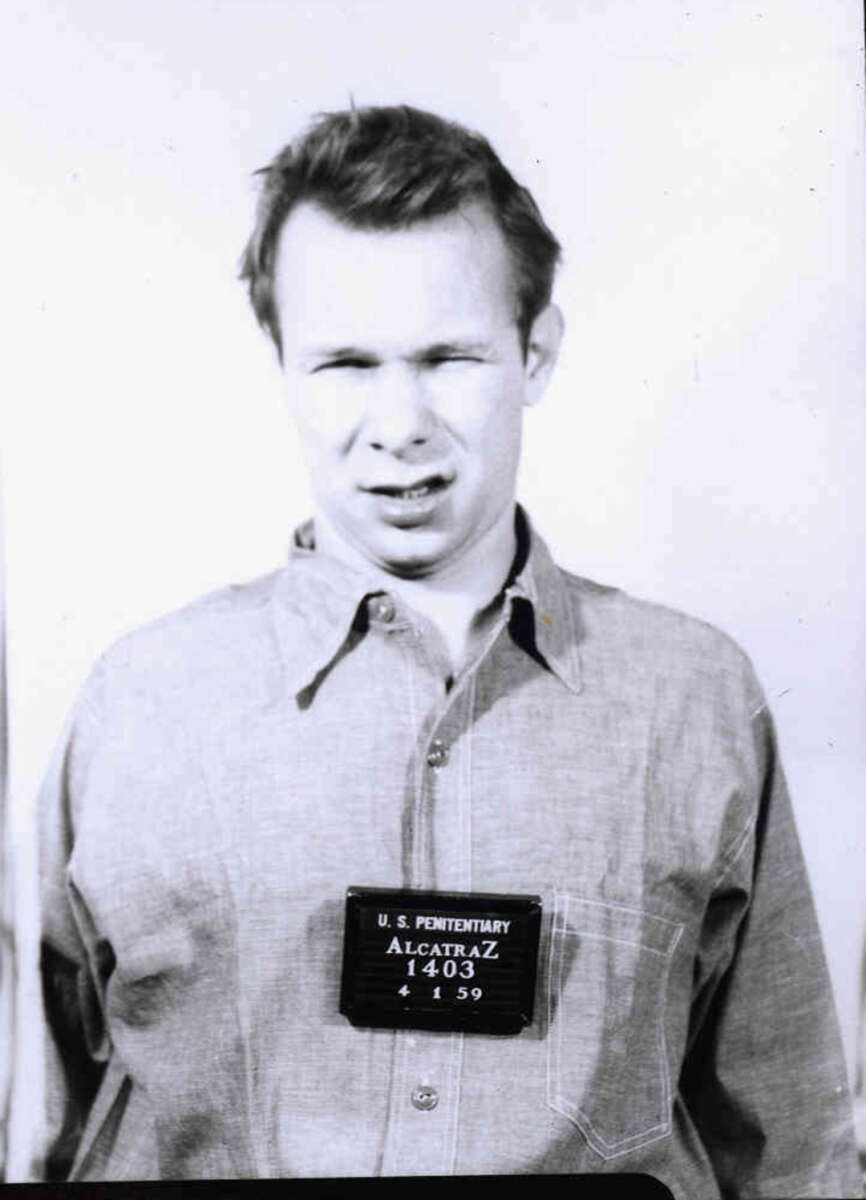 Black-and-white mugshot of a man in a button-up shirt with a sign reading "U.S. Penitentiary Alcatraz 1403 4 4 59" hanging on his chest. The man is looking slightly to the side.