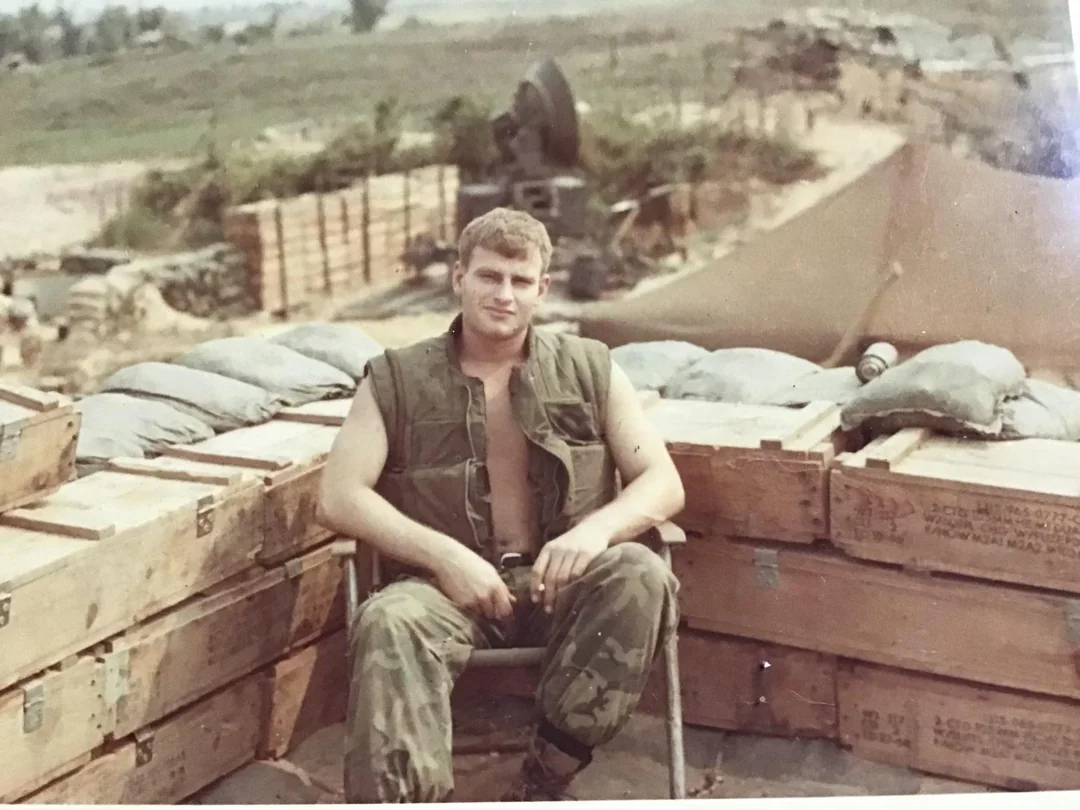 A young man in military fatigues sits relaxed on a chair among stacked wooden crates and sandbags in an outdoor camp, with a large tent and equipment visible in the background.