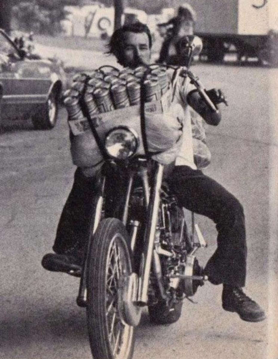 A man rides a motorcycle carrying a large bundle of canned drinks strapped to the handlebars, with one foot off the pedal, on a street with cars and another biker in the background.