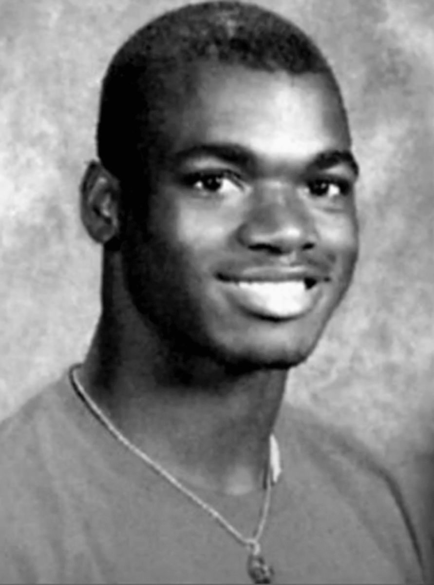 Black and white portrait of a young man smiling at the camera, wearing a necklace and a plain t-shirt, with a neutral textured background.