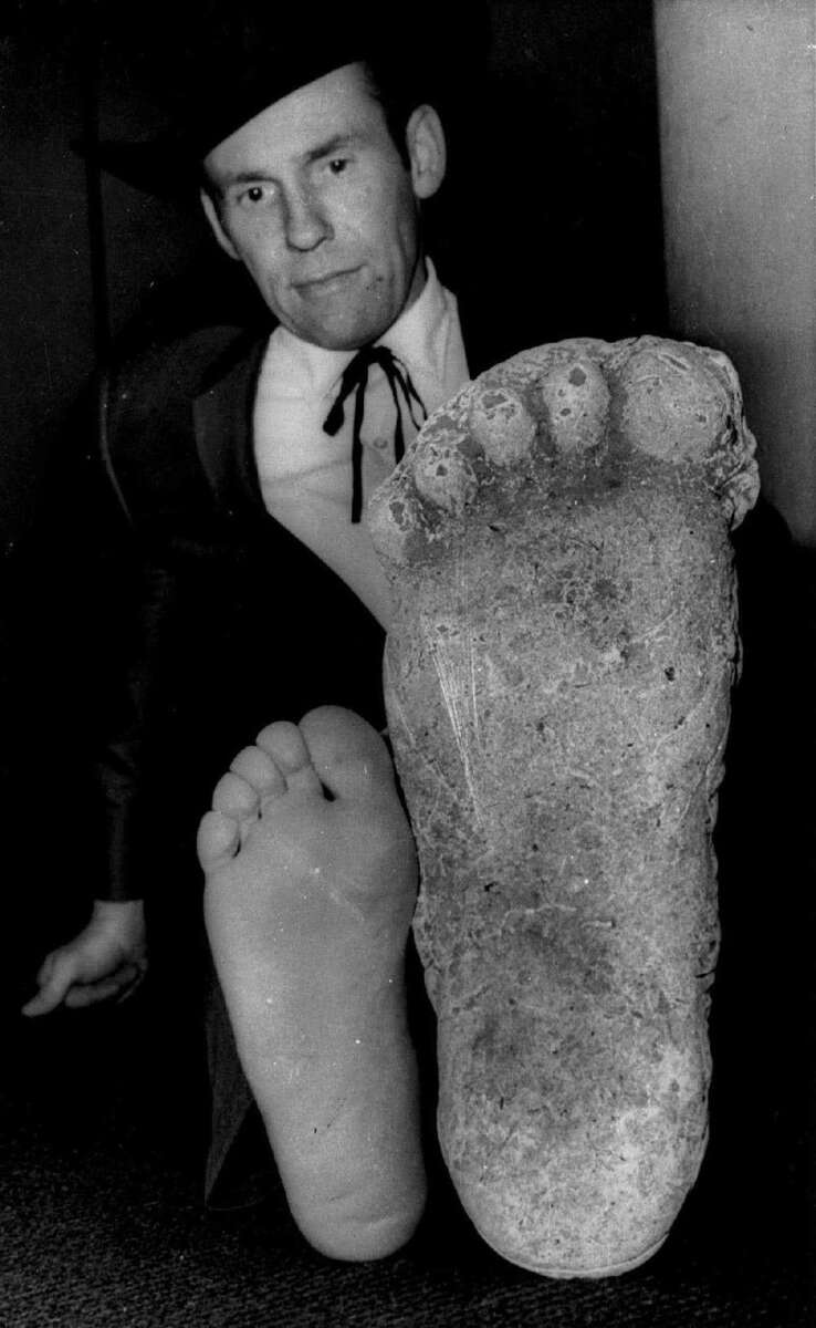 A man in a suit and hat sits on the floor, holding his bare foot next to a large plaster cast of an oversized footprint, highlighting the significant difference in size.