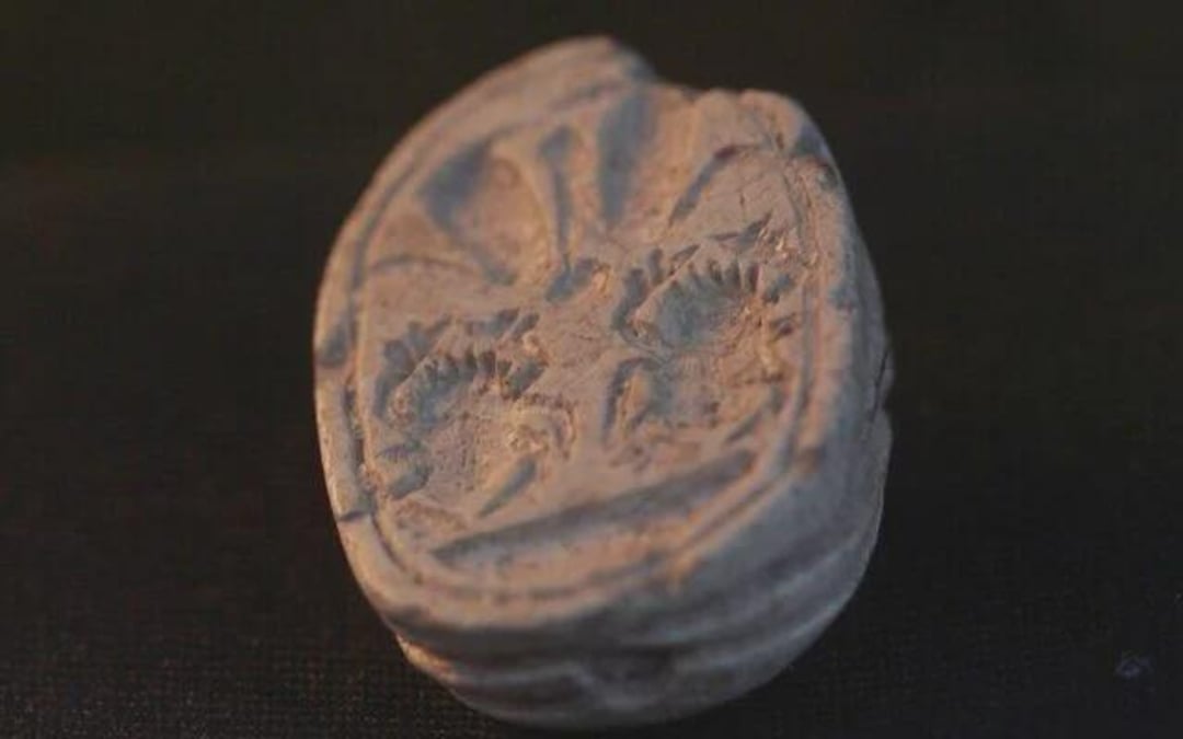 A close-up of an ancient clay seal impression with intricate, worn designs, shown on a dark background. The seal appears oval and features faded patterns, possibly depicting animals or symbols.