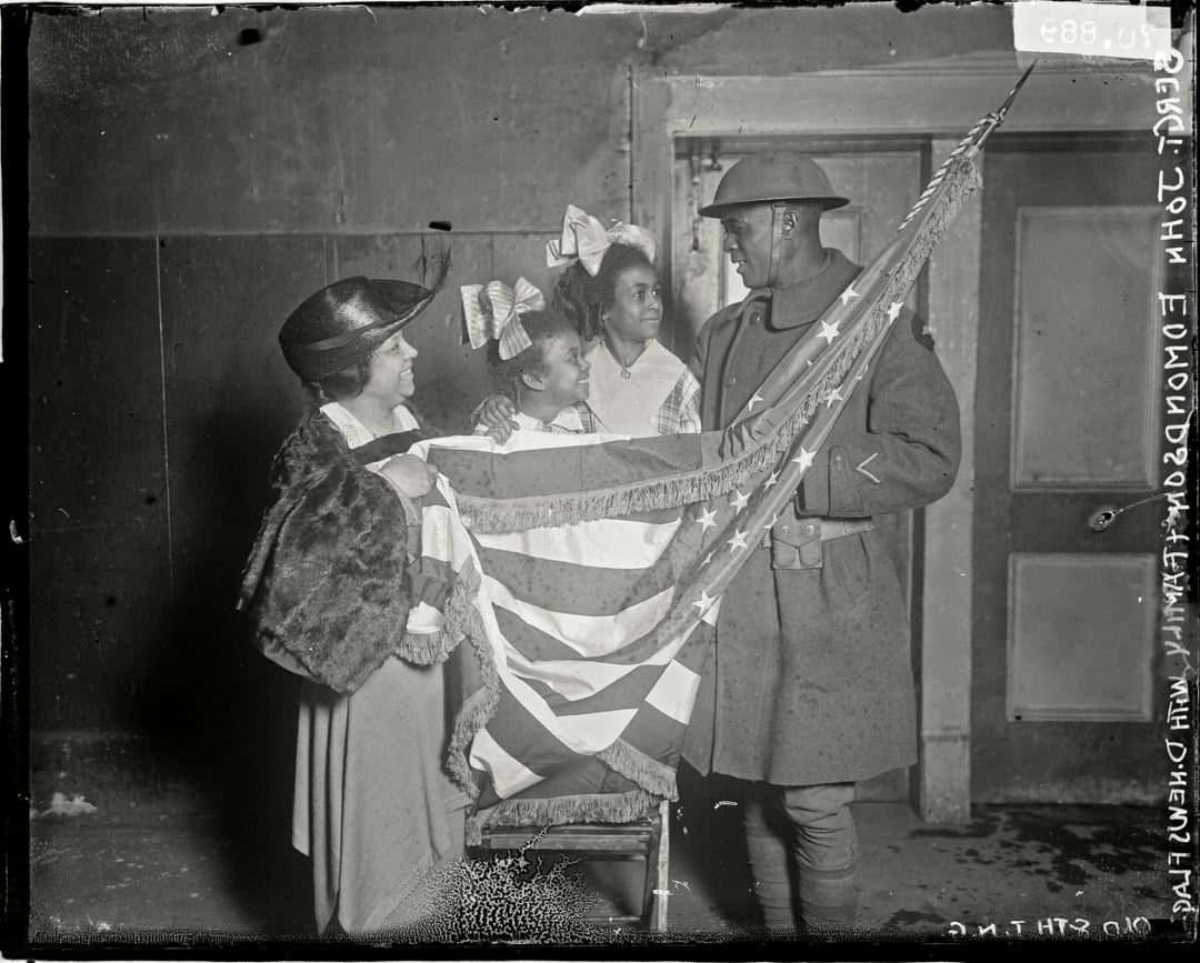 A Black World War I soldier in uniform stands holding an American flag with three smiling Black women and girls, one wearing a fur stole, in an indoor setting with plain walls and a door in the background.