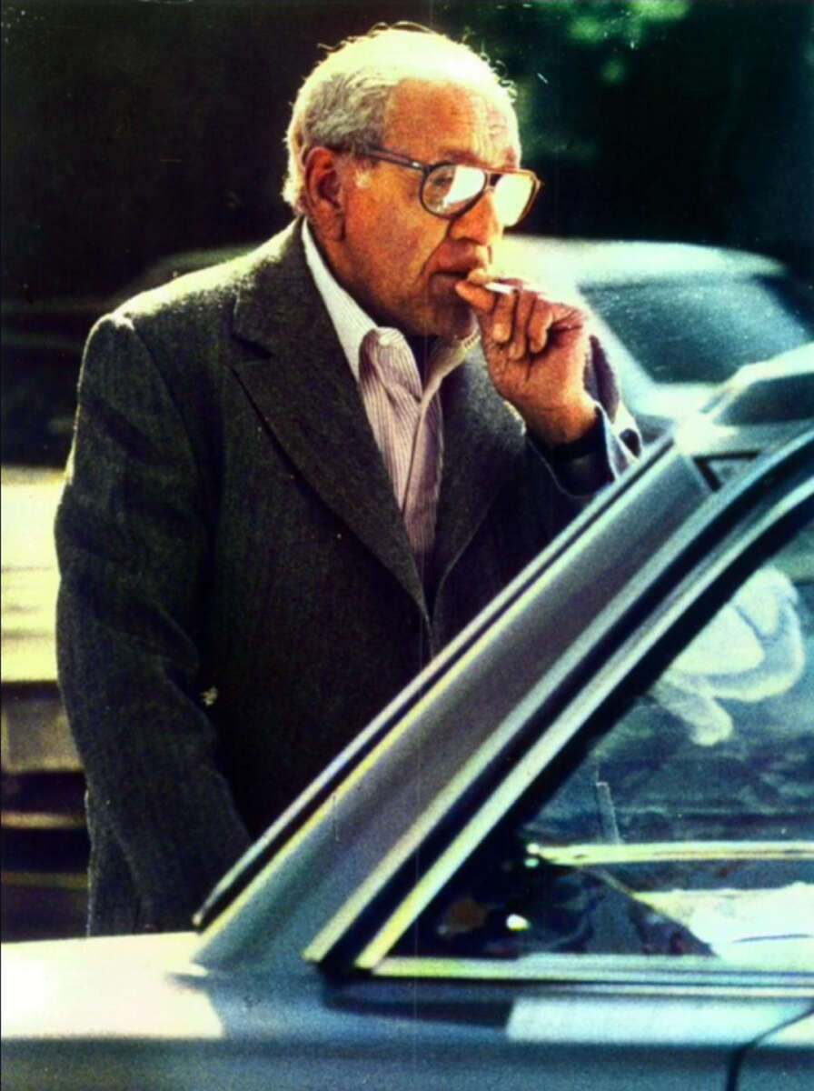 An elderly man with gray hair and glasses, wearing a suit jacket, smokes a cigarette while standing next to a car. The background is blurred, showing some daylight and parked cars.