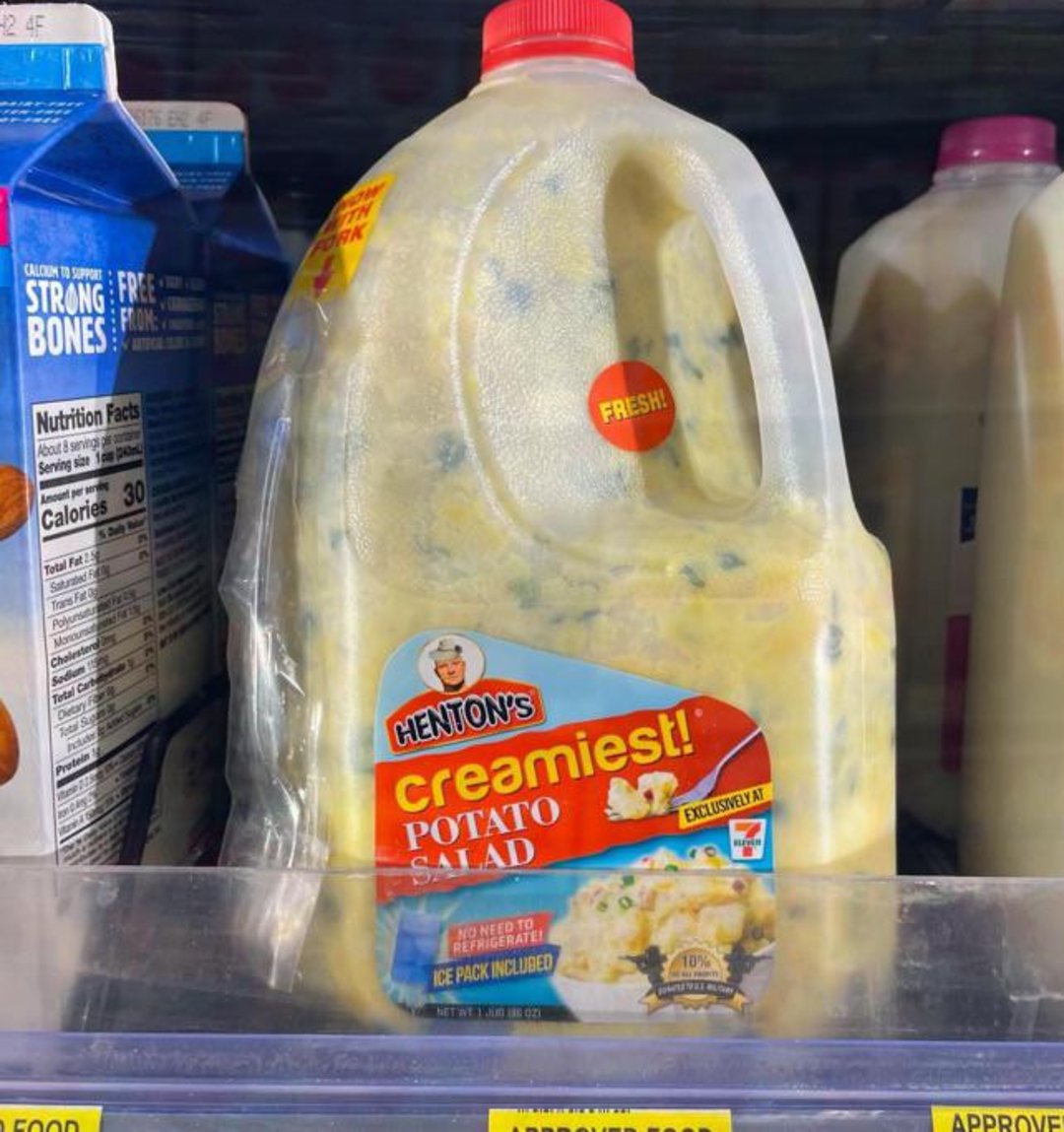 A large plastic jug labeled "Henton's Creamiest Potato Salad" sits on a grocery store shelf, next to cartons of milk. The jug has a red cap and a yellow label with product details.