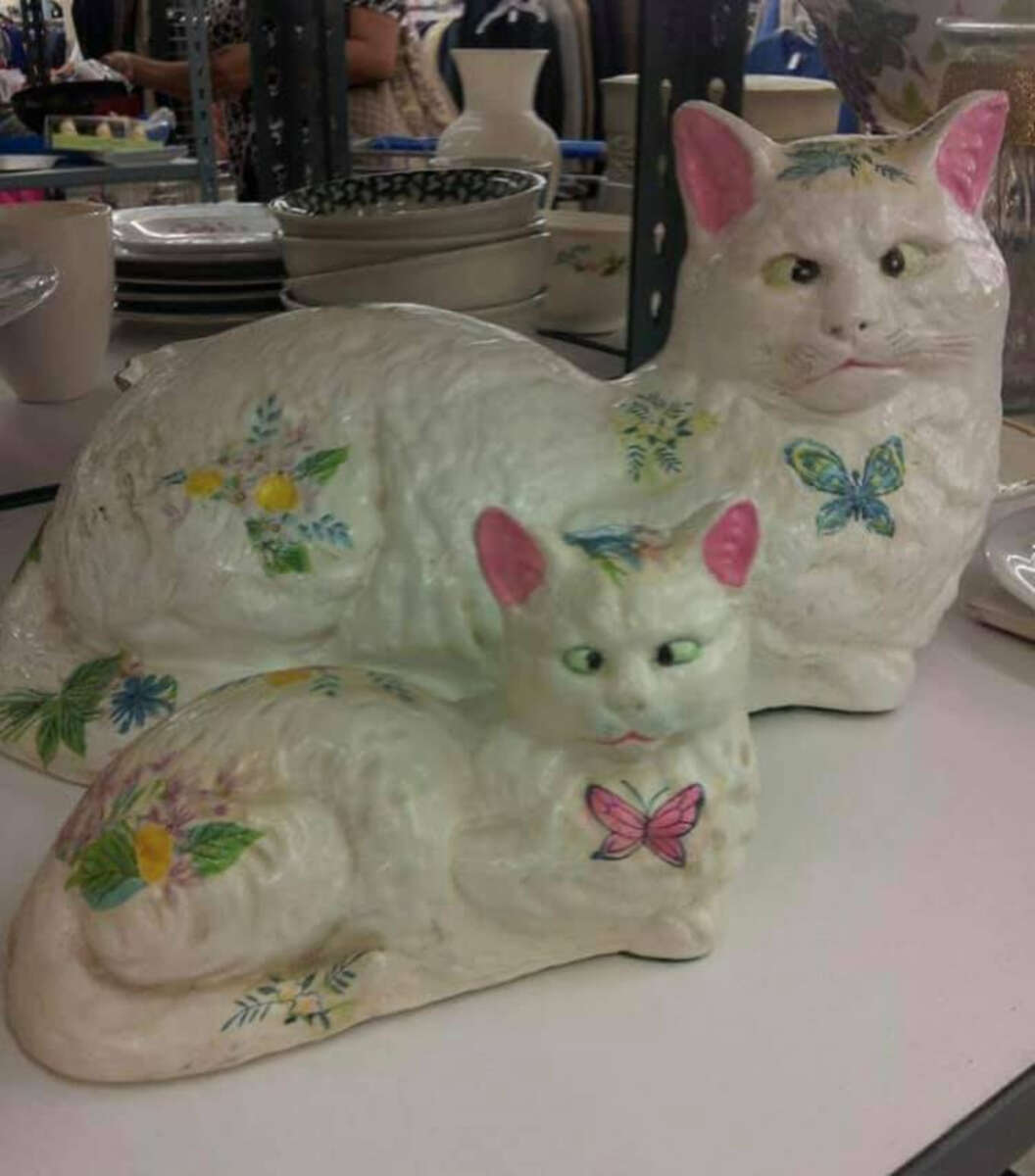 Two decorative ceramic cats with painted pastel flowers and butterflies sit on display in a shop; the larger cat is behind the smaller one, both white with pink ears and green eyes.