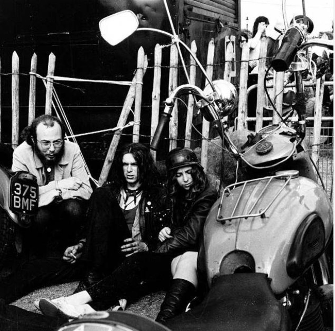 Three people sit on the ground beside a parked motorcycle, near a wooden fence. Two men and one woman look relaxed, wearing casual and leather clothing, with another person partly visible in the background.