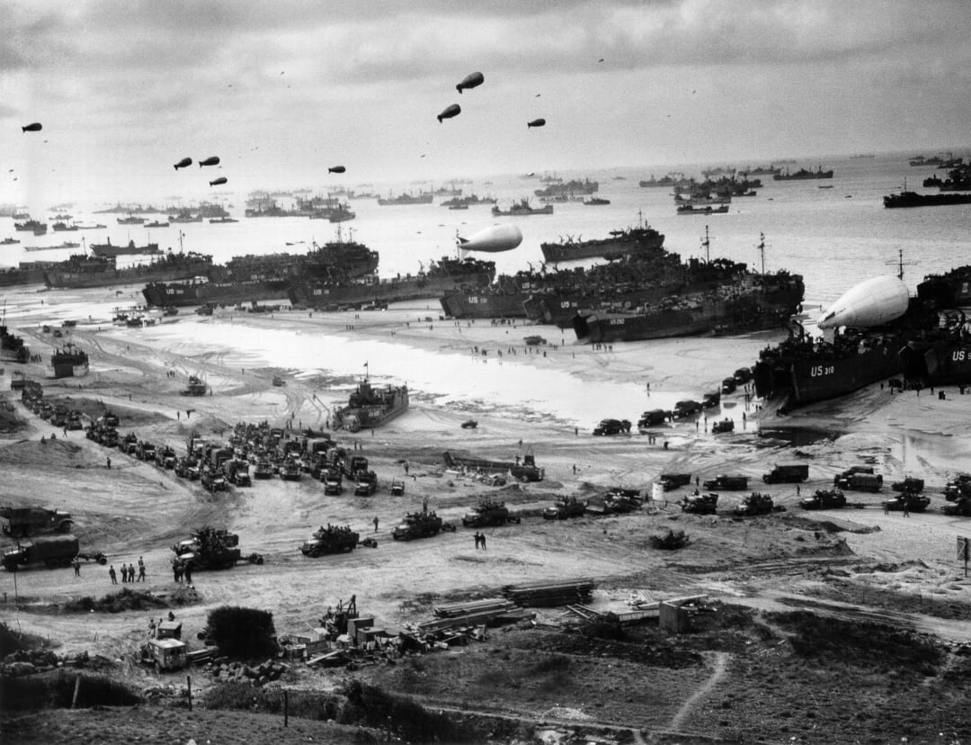 Black-and-white photo of D-Day landings: numerous ships, landing craft, and vehicles crowd the beach, with troops, tanks, and equipment coming ashore, while barrage balloons float in the sky above the scene.