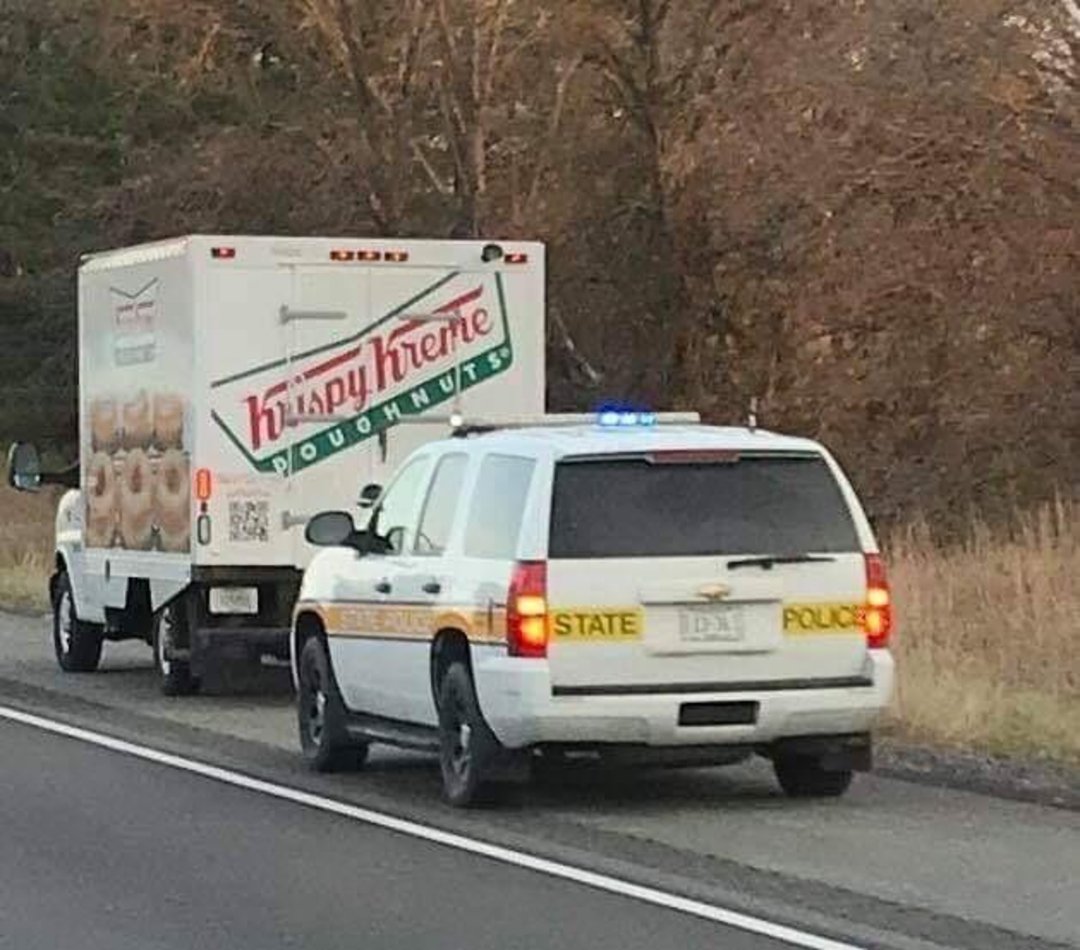 A Krispy Kreme doughnut truck is pulled over on the side of a road by a marked state police SUV with its lights on. Trees without leaves line the background.