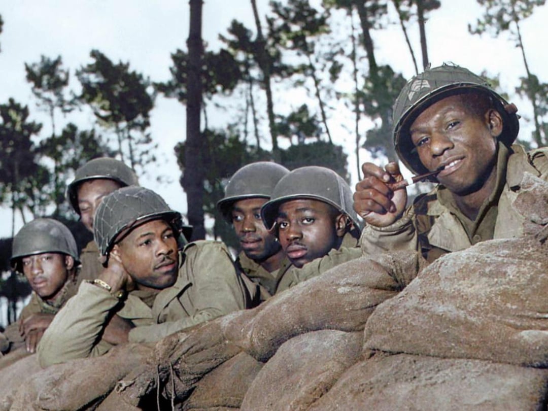 A group of African American soldiers in World War II uniforms crouch behind sandbags, some wearing helmets, with trees in the background. One soldier in front smiles and holds a pipe.
