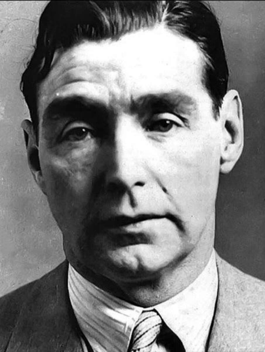 Black and white portrait of a middle-aged man with dark, wavy hair, wearing a suit, collared shirt, and tie. He has a neutral expression and is looking directly at the camera.