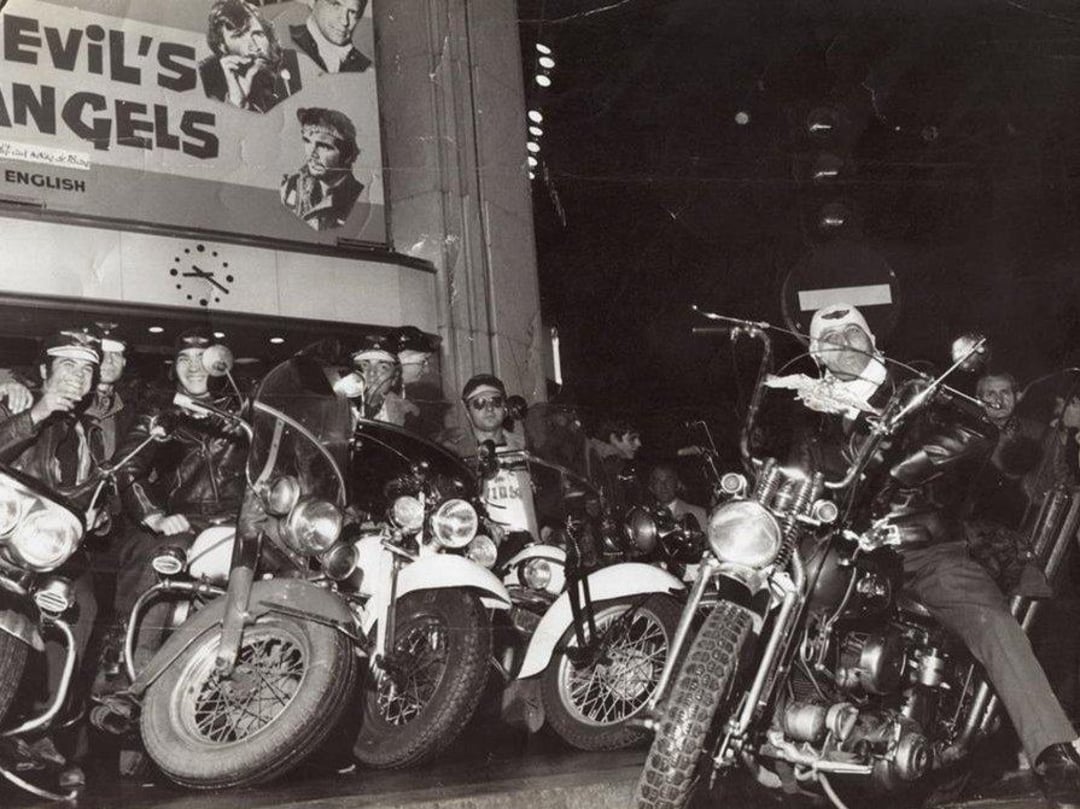 A group of bikers in leather jackets and sunglasses gather outside a theater showing the film "Devil’s Angels." Several motorcycles are parked in front, and a few bikers look toward the camera under the marquee.