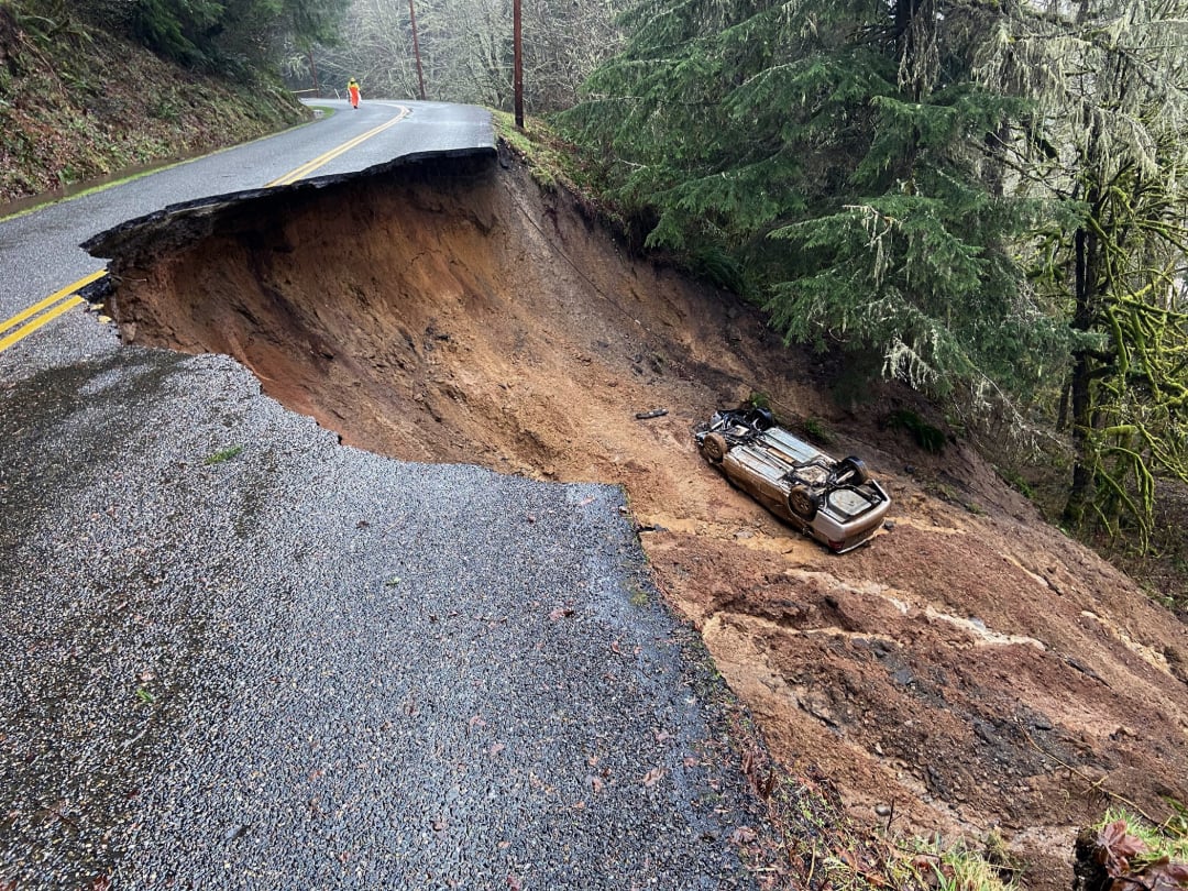 A large section of a road has collapsed in a landslide, leaving a deep, muddy hole with an overturned car at the bottom. A person in a reflective vest stands near the edge, observing the damage.