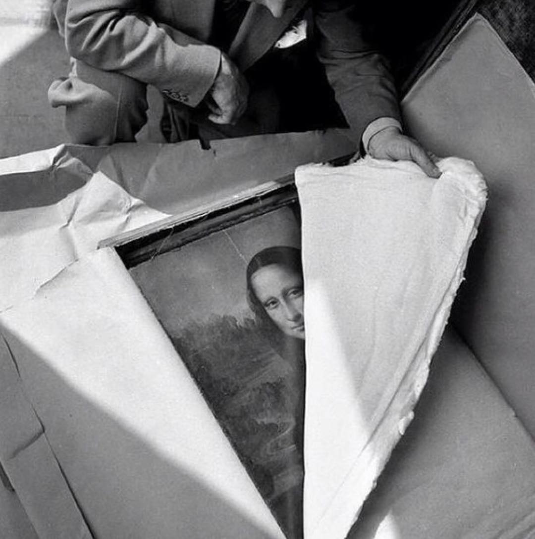 A person carefully unwraps a framed painting of the Mona Lisa, partially covered by protective paper, revealing her face while holding the edge of the wrapping.