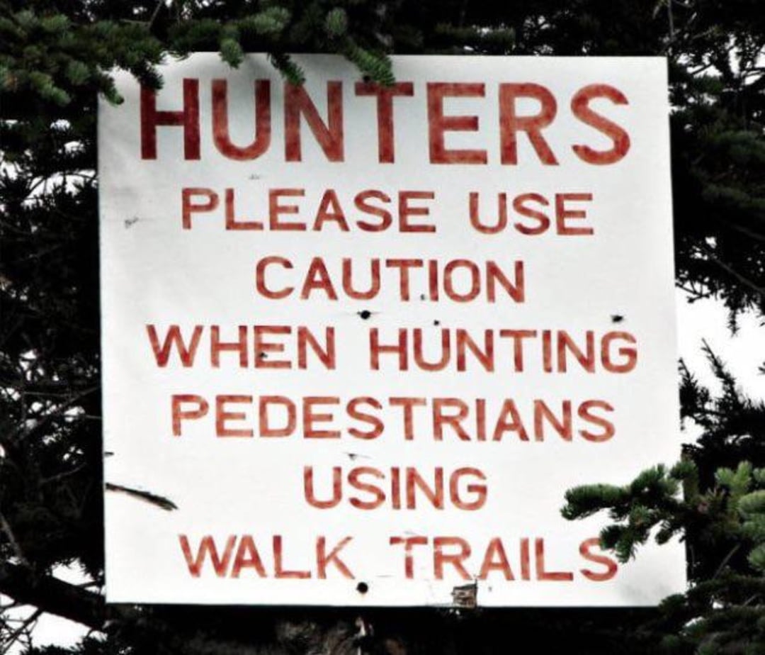 A sign on a tree reads: "HUNTERS PLEASE USE CAUTION WHEN HUNTING PEDESTRIANS USING WALK TRAILS," with green branches partially covering the bottom corners.