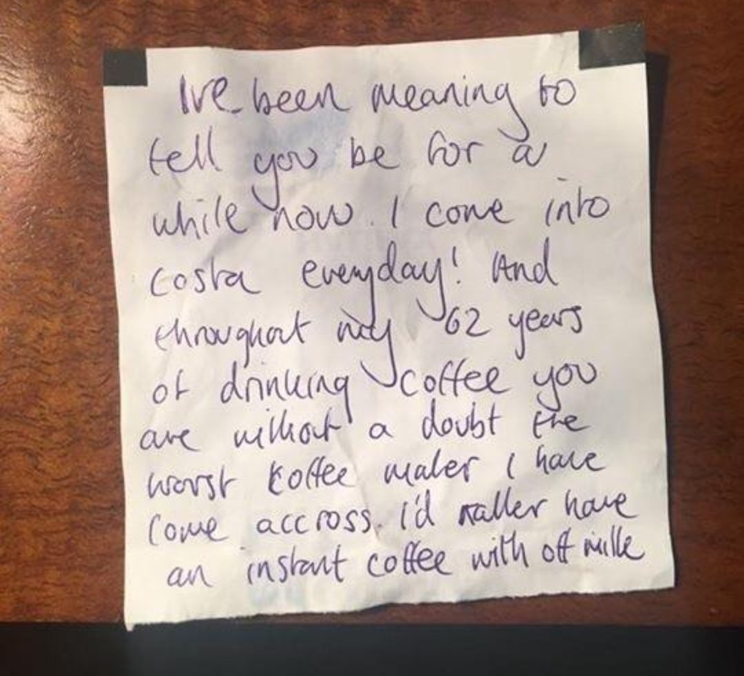 A handwritten note on white paper reads: “I've been meaning to tell you for a while now: I come into Costa everyday! And throughout my 62 years of drinking coffee you are without a doubt the worst coffee maker I have come across. I’d rather have an instant coffee with off milk.” The note is taped to a brown wooden surface.