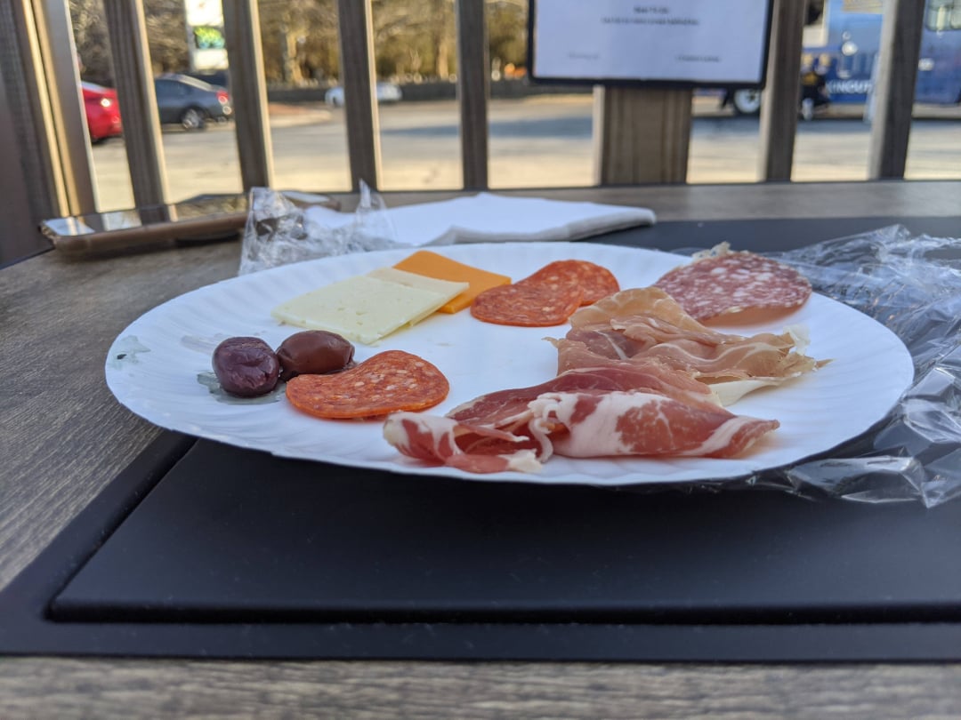A white paper plate on a wooden table holds slices of cheese, salami, prosciutto, two olives, and pepperoni. The plate is outdoors with cars and trees visible in the background.