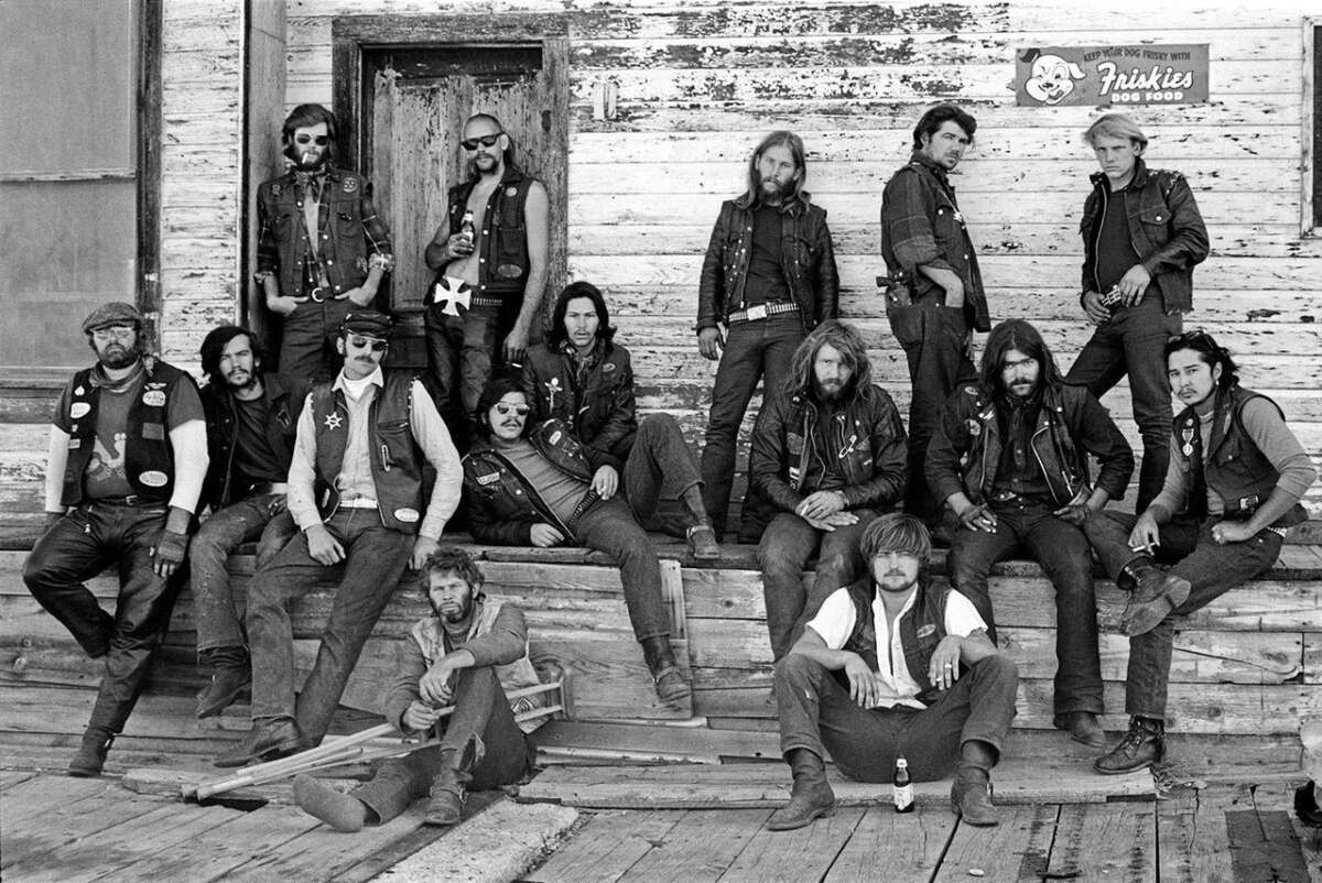 A group of men in biker vests pose in front of a weathered wooden building, some standing, some sitting on the porch. The mood is serious, and the setting has a rugged, vintage look.