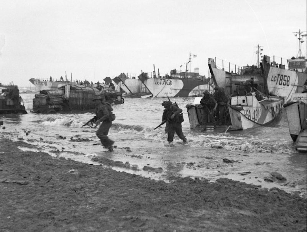 Soldiers disembark from landing crafts onto a beach during a World War II military operation, with vehicles and additional troops following behind in shallow waters under a cloudy sky.