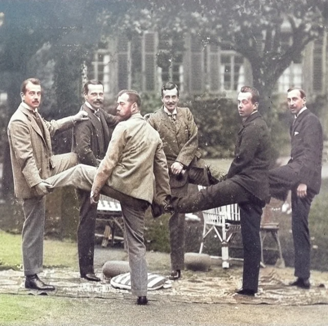 Six men in vintage suits stand in a row outdoors, each with one leg lifted and resting on the knee of the man beside him. They appear to be posing playfully in front of a large building and garden furniture.
