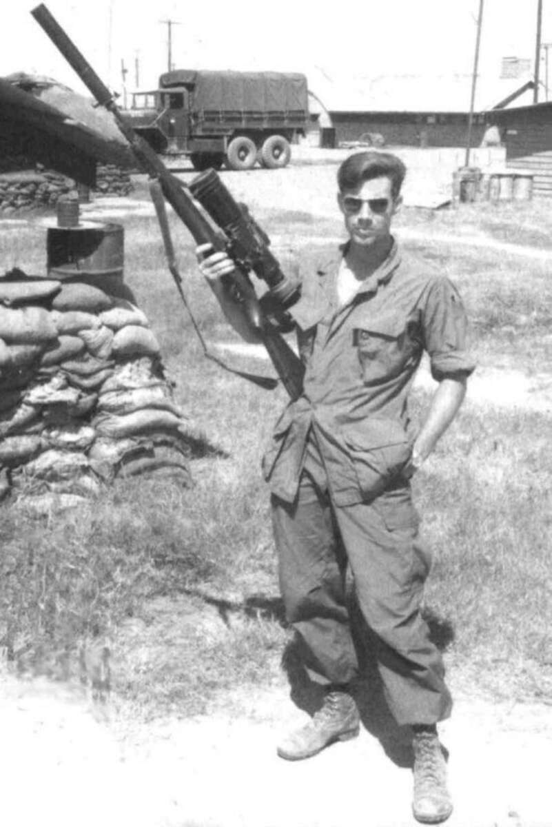 A soldier in military fatigues stands outdoors holding a rifle upright over his shoulder. He wears sunglasses and boots, with sandbags and military vehicles visible in the background.