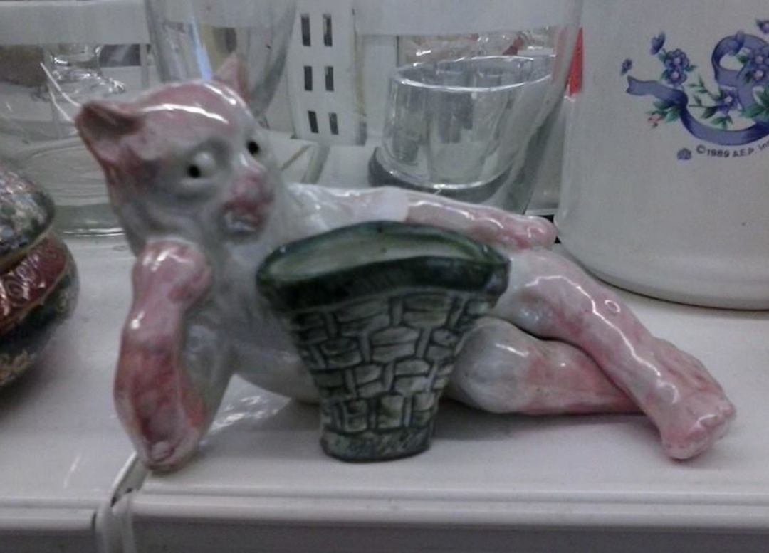 A small ceramic figurine of a lounging pink and white cat with a surprised expression, leaning on a stone-patterned object, displayed on a white shelf among glassware and other ceramics.
