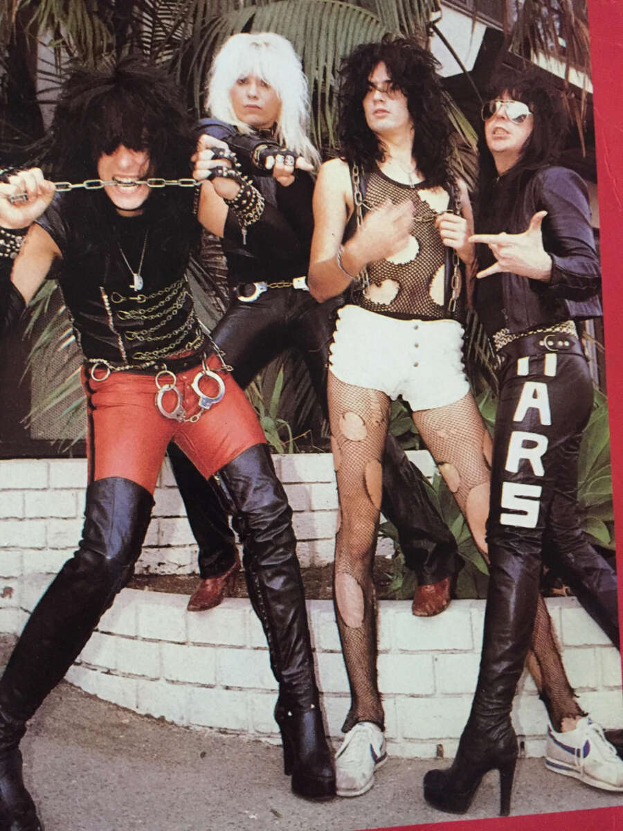 Four people dressed in edgy, glam rock outfits with dramatic hairstyles pose outdoors in front of a brick planter and foliage. Their clothing includes leather, fishnet, studs, chains, and large letters spelling “STARS” on one leg.