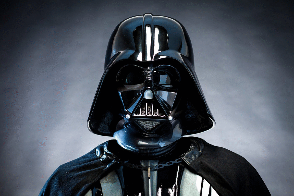 A person wearing a shiny black Darth Vader helmet and cape stands against a neutral gray background, facing forward. The helmet reflects light and the costume includes molded black armor.