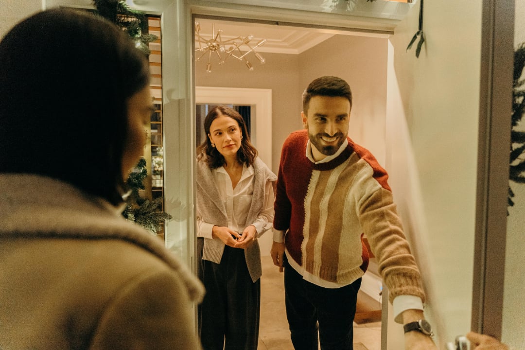 A man and woman stand at an open door, smiling warmly as they enter a house, greeted by a person whose back is to the camera. The scene is cozy, suggesting a friendly holiday or winter gathering.