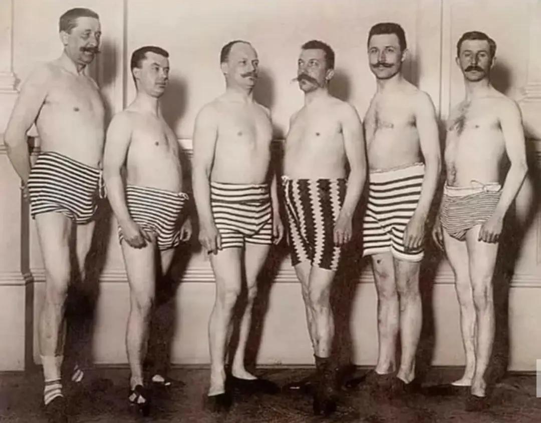 Six men pose indoors in vintage, striped swimsuits from the early 1900s. They stand in a row against a light-colored wall, with most of them sporting mustaches and looking toward the camera.
