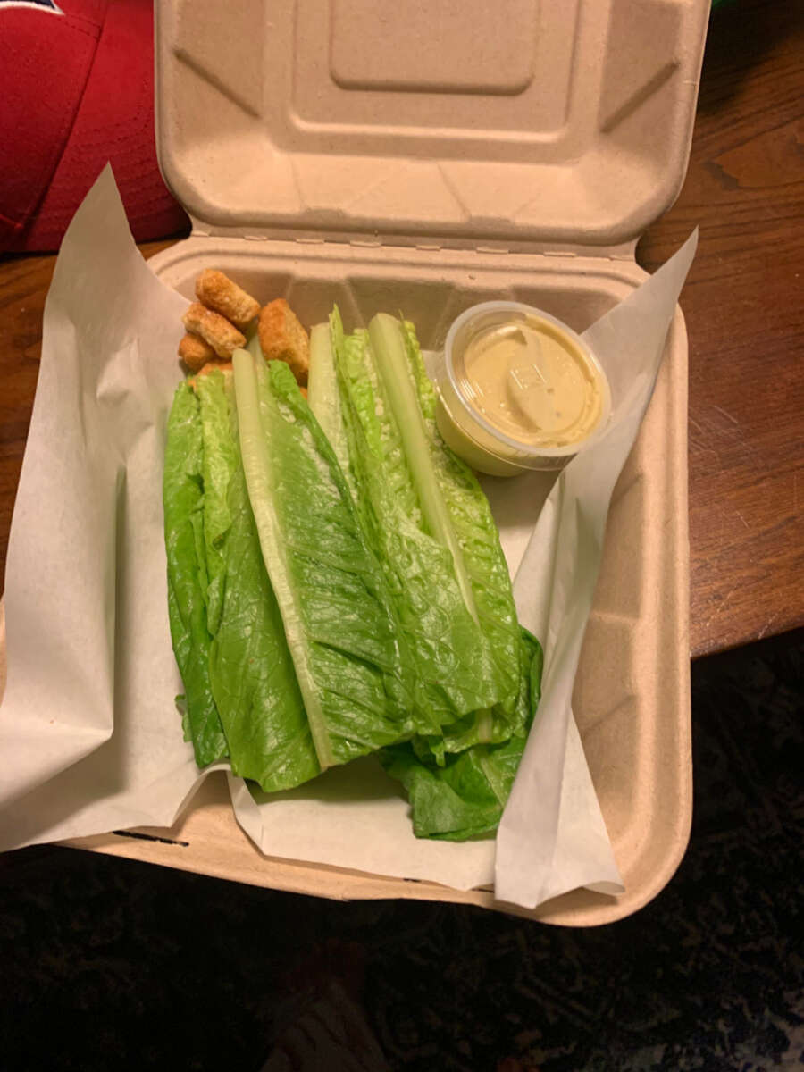 A takeout box with romaine lettuce leaves, a few croutons, a cup of creamy dressing, and no visible protein or other salad toppings, placed on a wooden table.