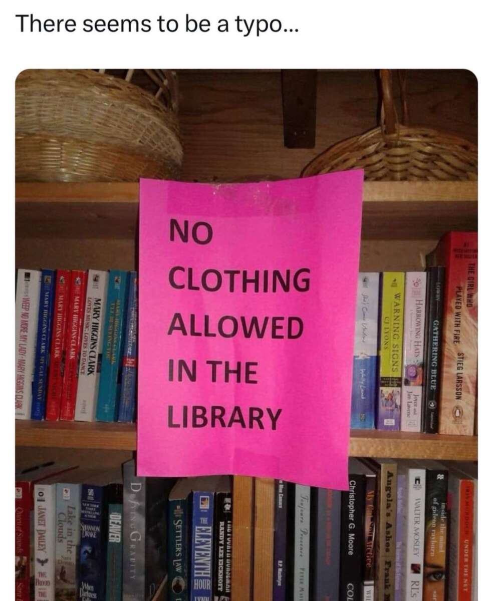 A bright pink sign on a bookshelf reads, “NO CLOTHING ALLOWED IN THE LIBRARY.” The text appears to be an accidental typo, and the sign is surrounded by books and wicker baskets.
