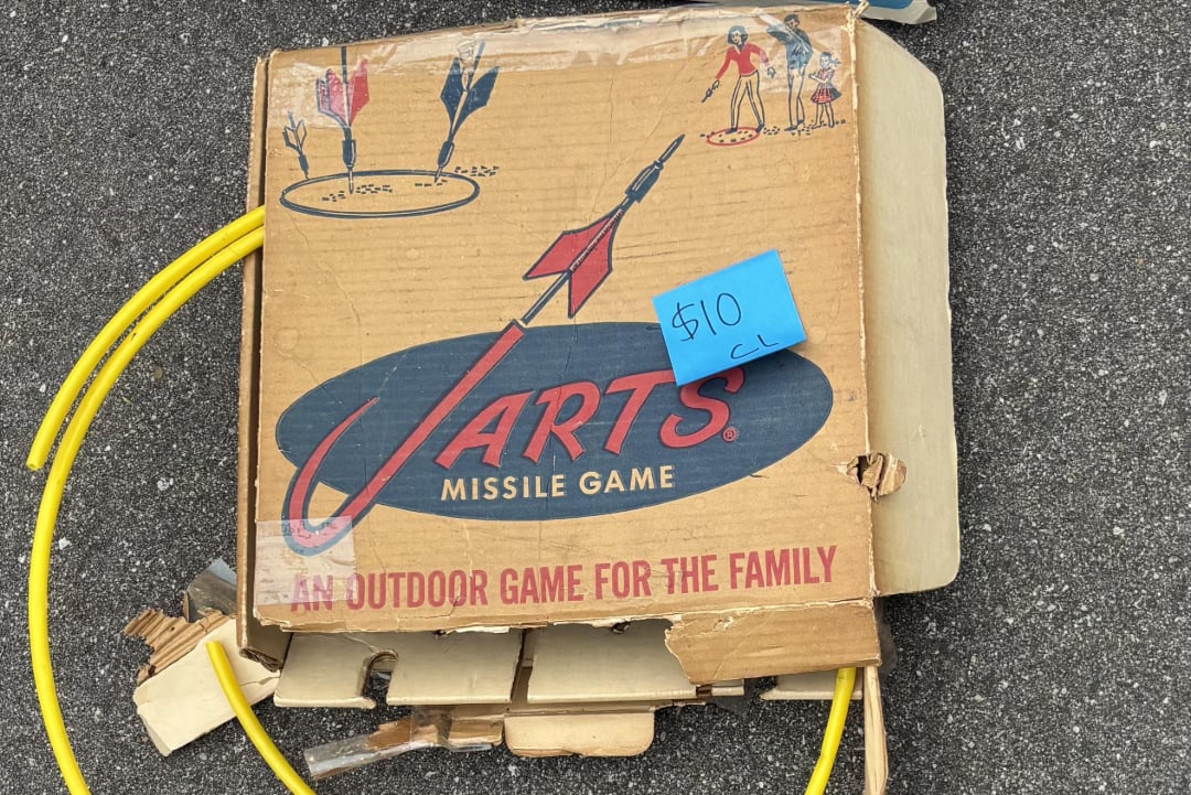 A worn cardboard box labeled "Jarts Missile Game: An Outdoor Game for the Family" sits on pavement, with yellow plastic rings and a $10 price tag attached. The box features drawings of people playing the game.