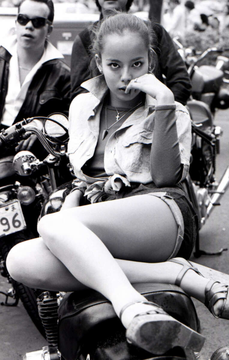 A young woman sits on a motorcycle with one leg crossed, resting her hand near her mouth and gazing intently at the camera. She wears denim shorts, a tied shirt, and sandals; people and motorcycles are visible in the background.