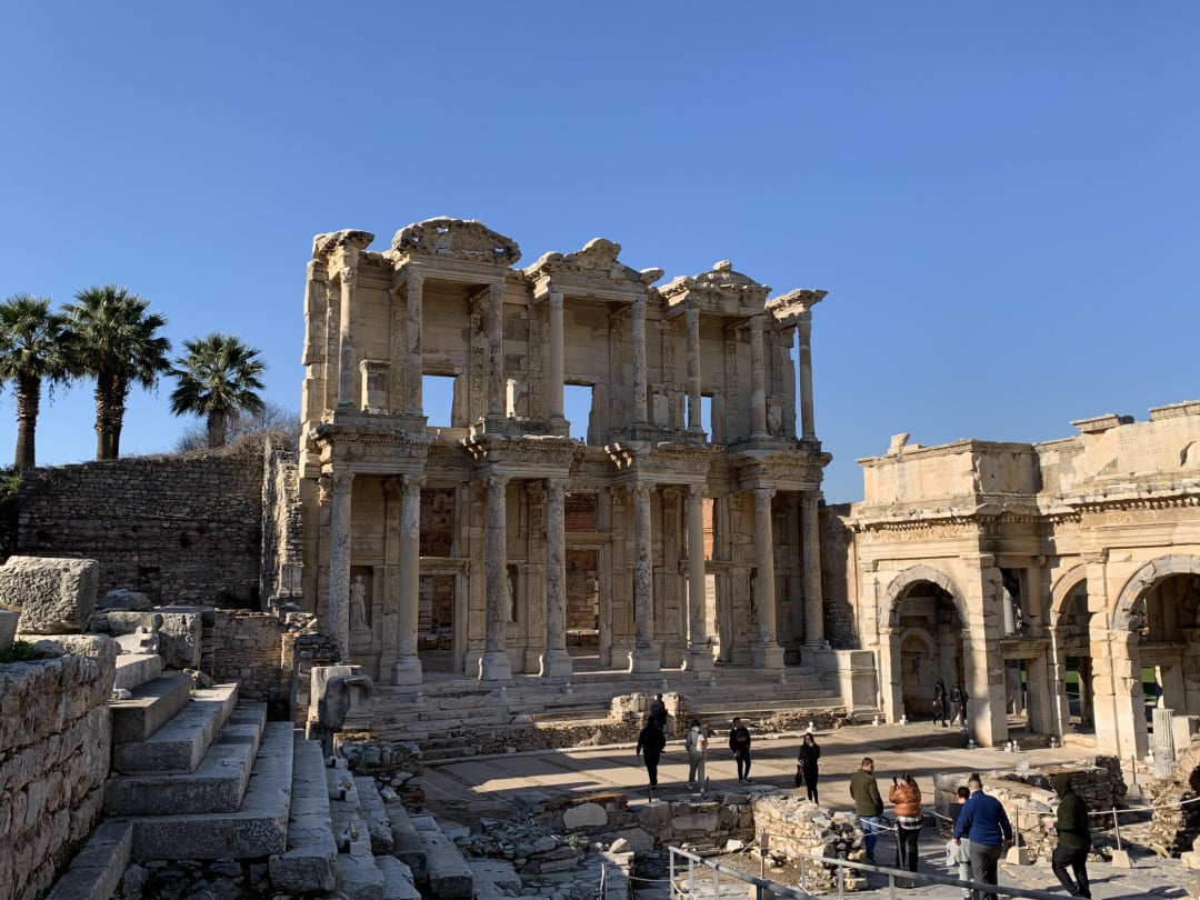 Ancient ruins of the Library of Celsus in Ephesus, Turkey, with a clear blue sky, a few people exploring the site, palm trees to the left, and stone arches and steps in the foreground.