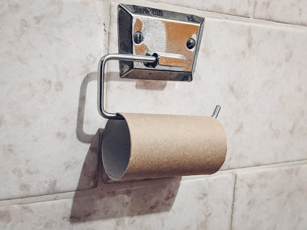 An empty cardboard toilet paper roll is left on a metal holder attached to a tiled bathroom wall. The roll's paper has been completely used up.