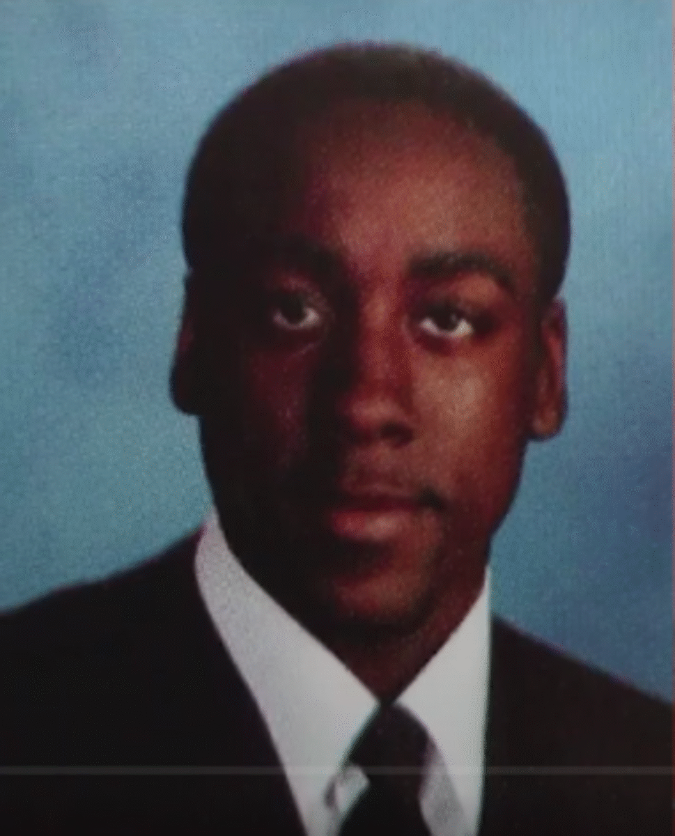 A young man wearing a dark suit, white shirt, and dark tie poses for a formal portrait against a blue background.