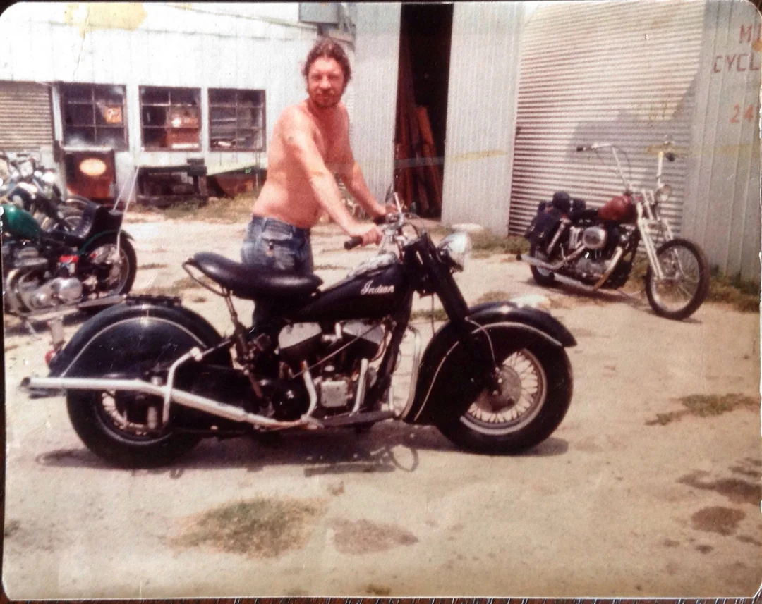 A shirtless man stands outdoors next to a vintage black Indian motorcycle. Several other motorcycles are parked nearby, and a corrugated metal building is in the background.