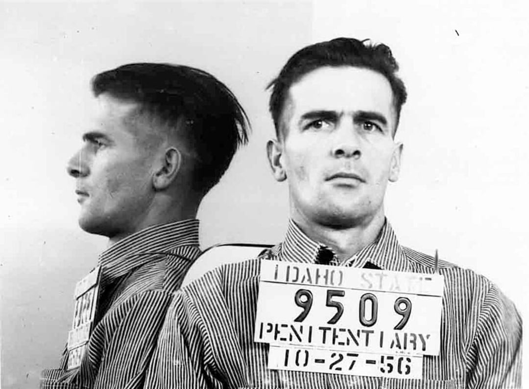 Black and white mugshot of a man with short dark hair, shown from the front and side, wearing a striped shirt and holding a sign reading “IDAHO STATE 9509 PENITENTIARY 10-27-56.”