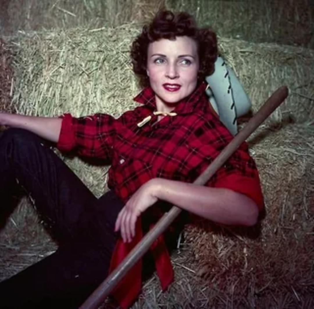 A woman in a red plaid shirt and dark pants leans against a hay bale, holding a pitchfork. She has short, curly hair and wears red lipstick, posed in a rustic, barn-like setting.