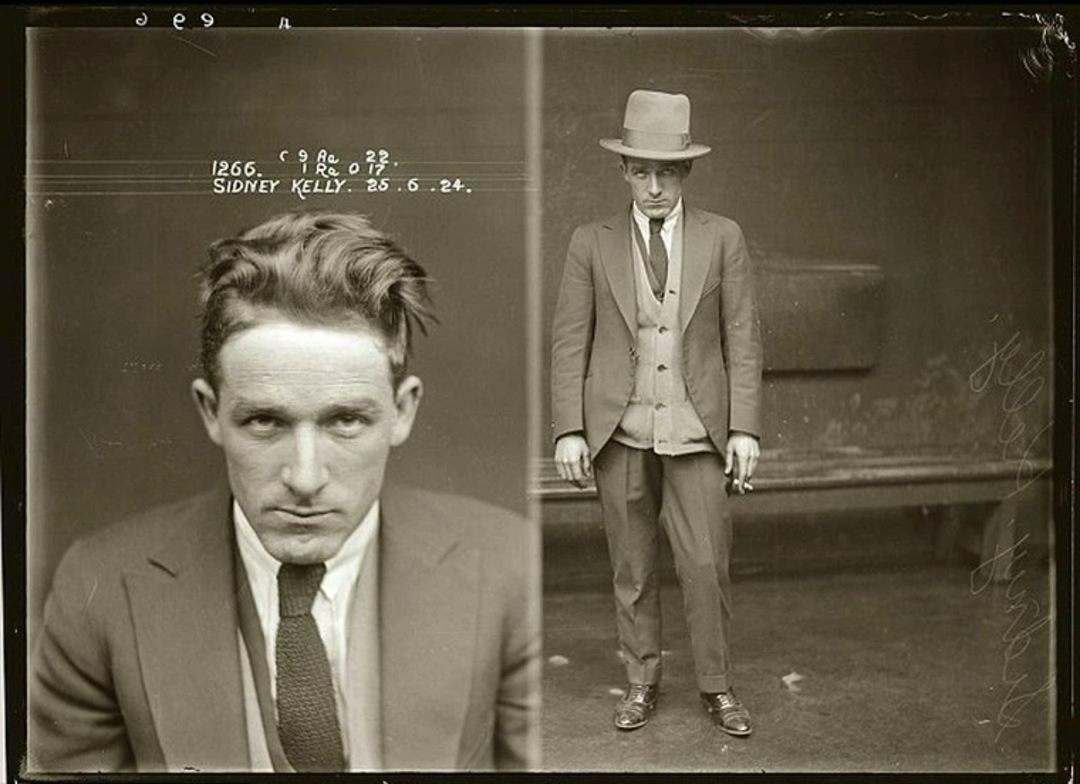 Black-and-white mugshot of a man in a suit and tie, with one close-up of his face and one full-body shot. He wears a hat in the full-body shot. Text above the left image reads "SIDNEY KELLY 25.6.24.