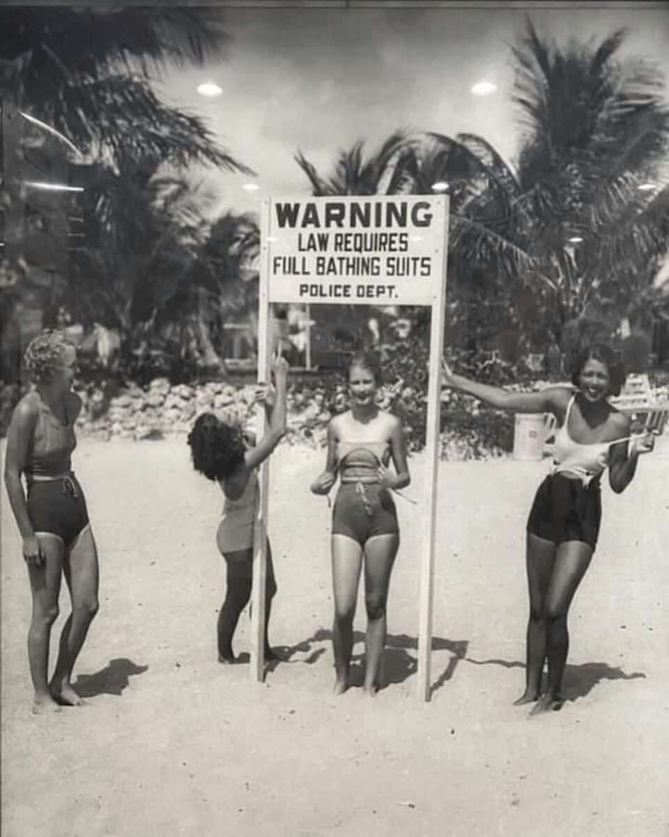 Four women in vintage swimsuits pose and smile by a beach sign that reads, "WARNING LAW REQUIRES FULL BATHING SUITS POLICE DEPT." Palm trees and sand appear in the background.