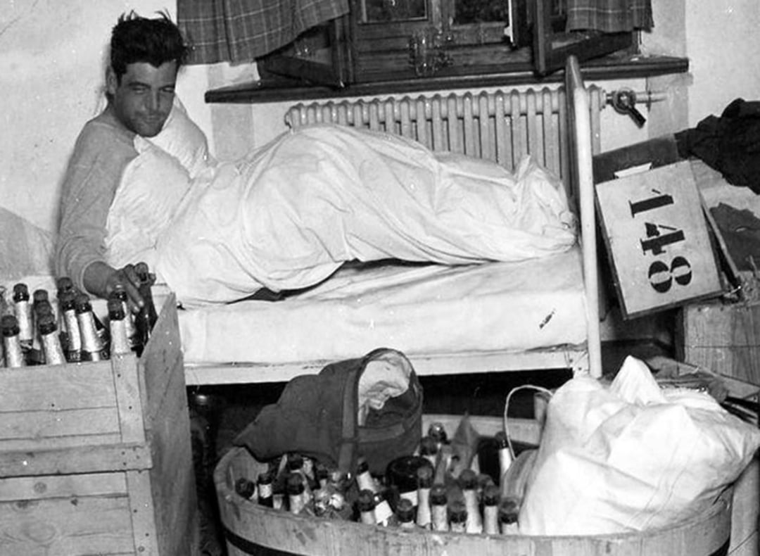 A man lies in bed, covered by a blanket, surrounded by crates and a tub filled with bottles. There is a large sign with the number 148 leaning against the bed, and a window with curtains in the background.