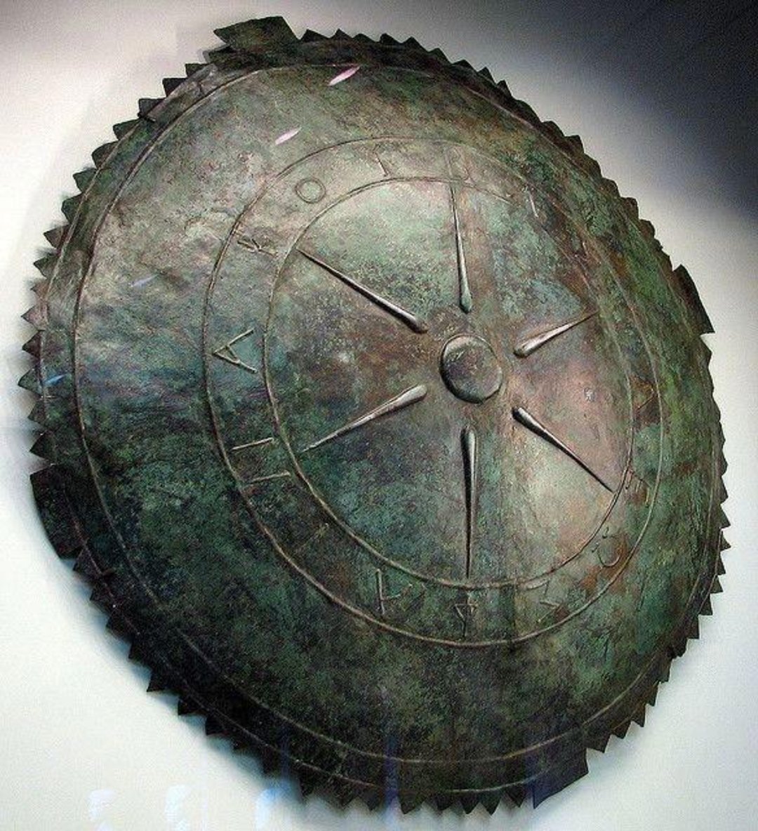 A large, round bronze shield with a worn, greenish patina, featuring a central star-like design and ancient markings or letters in a circular arrangement near the outer edge. The rim is edged with triangular notches.