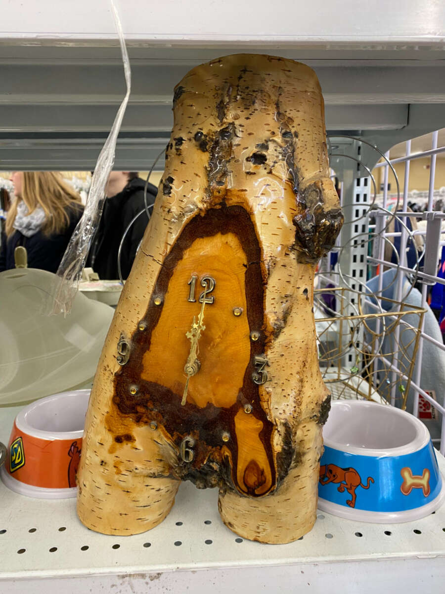 A handmade clock embedded in a piece of tree trunk sits on a shelf, surrounded by colorful pet bowls. People and various items are visible in the background.