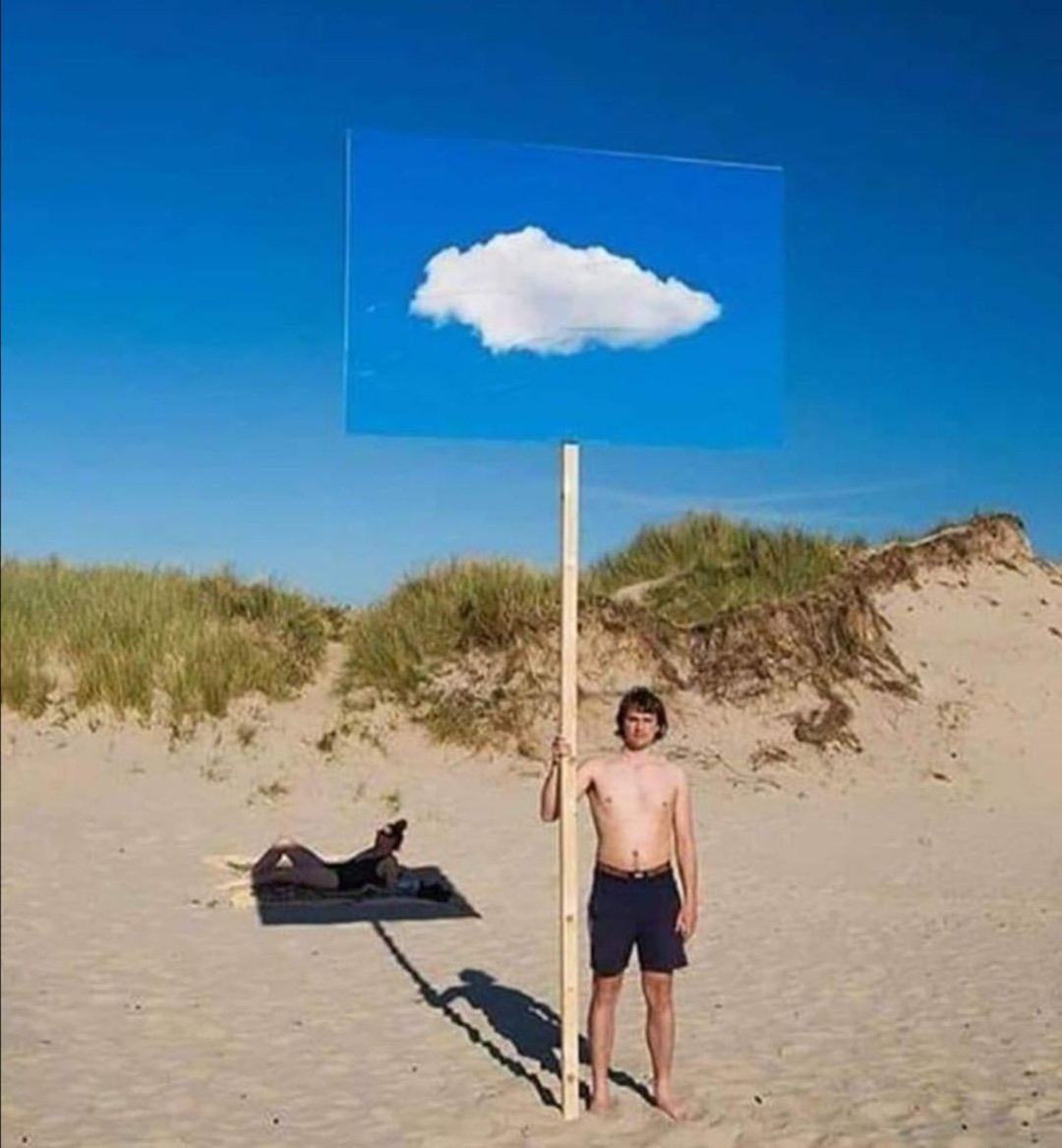 A shirtless person stands on a sandy beach holding a tall sign with a painted cloud that aligns perfectly with the sky, blending in with the real clouds. Grassy dunes and another person lying on a towel are in the background.