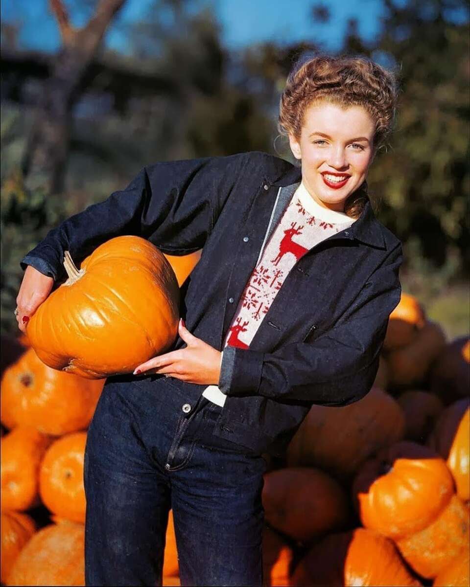 A smiling woman with curled hair holds a large pumpkin while standing in front of a pile of pumpkins. She wears a dark jacket, jeans, and a white sweater with red patterns. The background shows trees in sunlight.