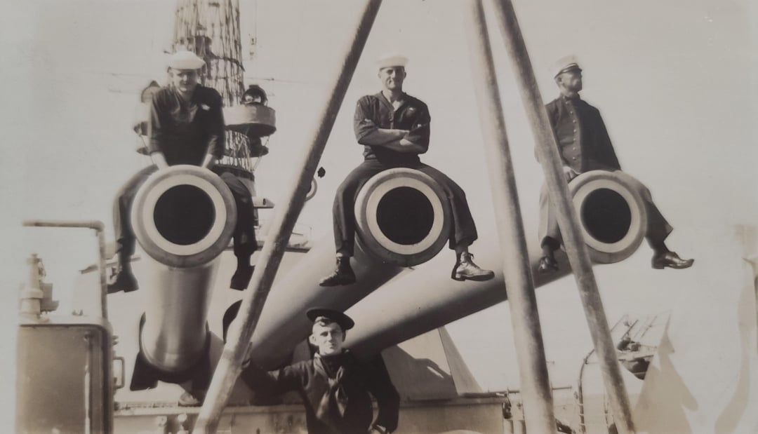 Four sailors in uniform pose on a battleship; three sit atop large naval gun barrels, while one stands below. The photo is black and white, and the scene appears to be from the early to mid-20th century.